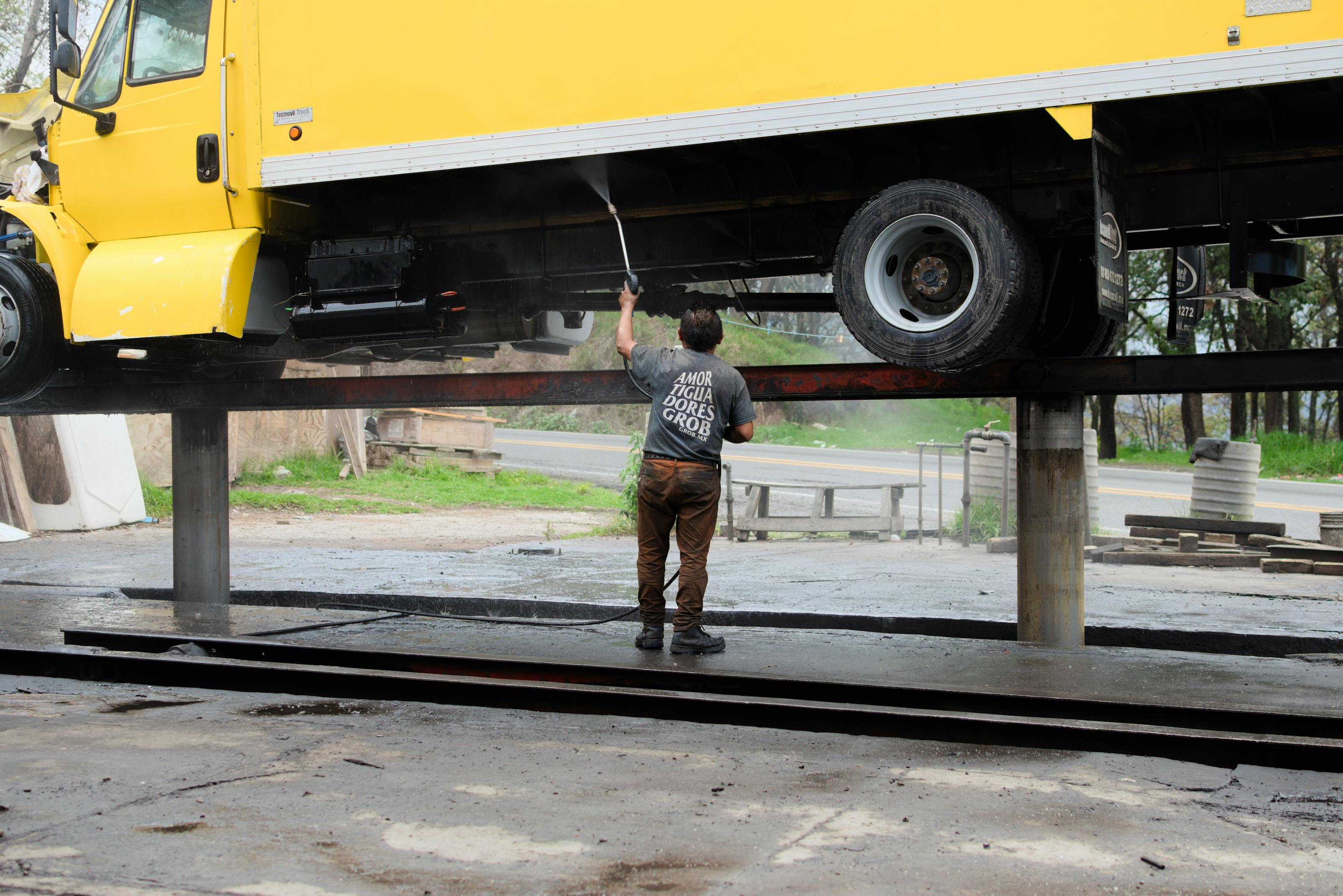 A pressure washing technician cleaning the underside of a raised yellow box truck at a fleet service yard - the kind of recurring commercial account that converts a residential pressure washing business into a route-density commercial operation in 2026.