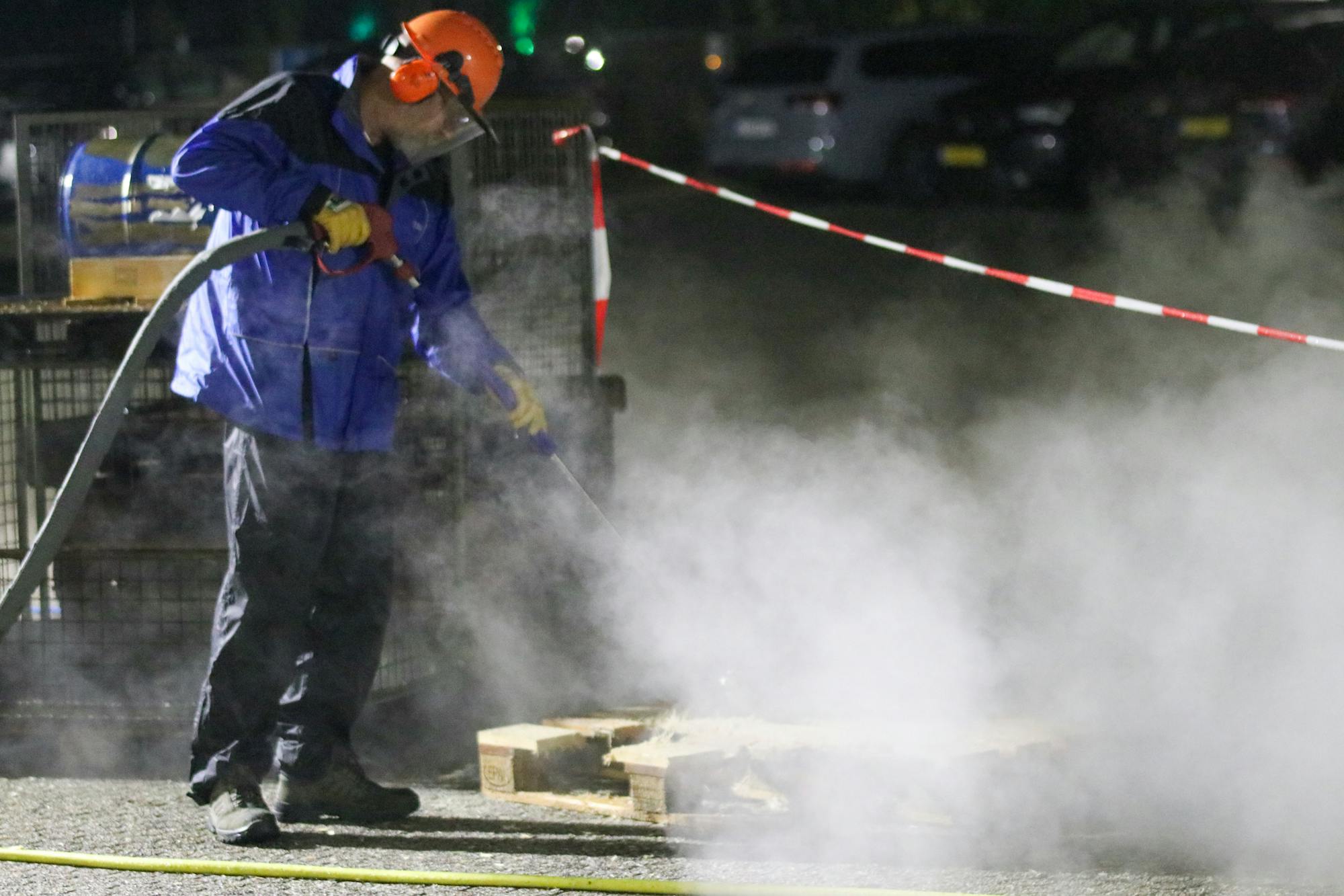 A pressure washing professional in full PPE (hard hat with face shield, chemical-resistant rain suit) working a high-pressure wand with steam visible - the kind of hot-water commercial cleaning work that runs 30-50% premium pricing over residential cold-water in 2026.